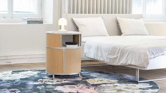 Rundes Nachttischmöbel Wogg No.4004 mit Holzfurnier neben einem Bett platziert