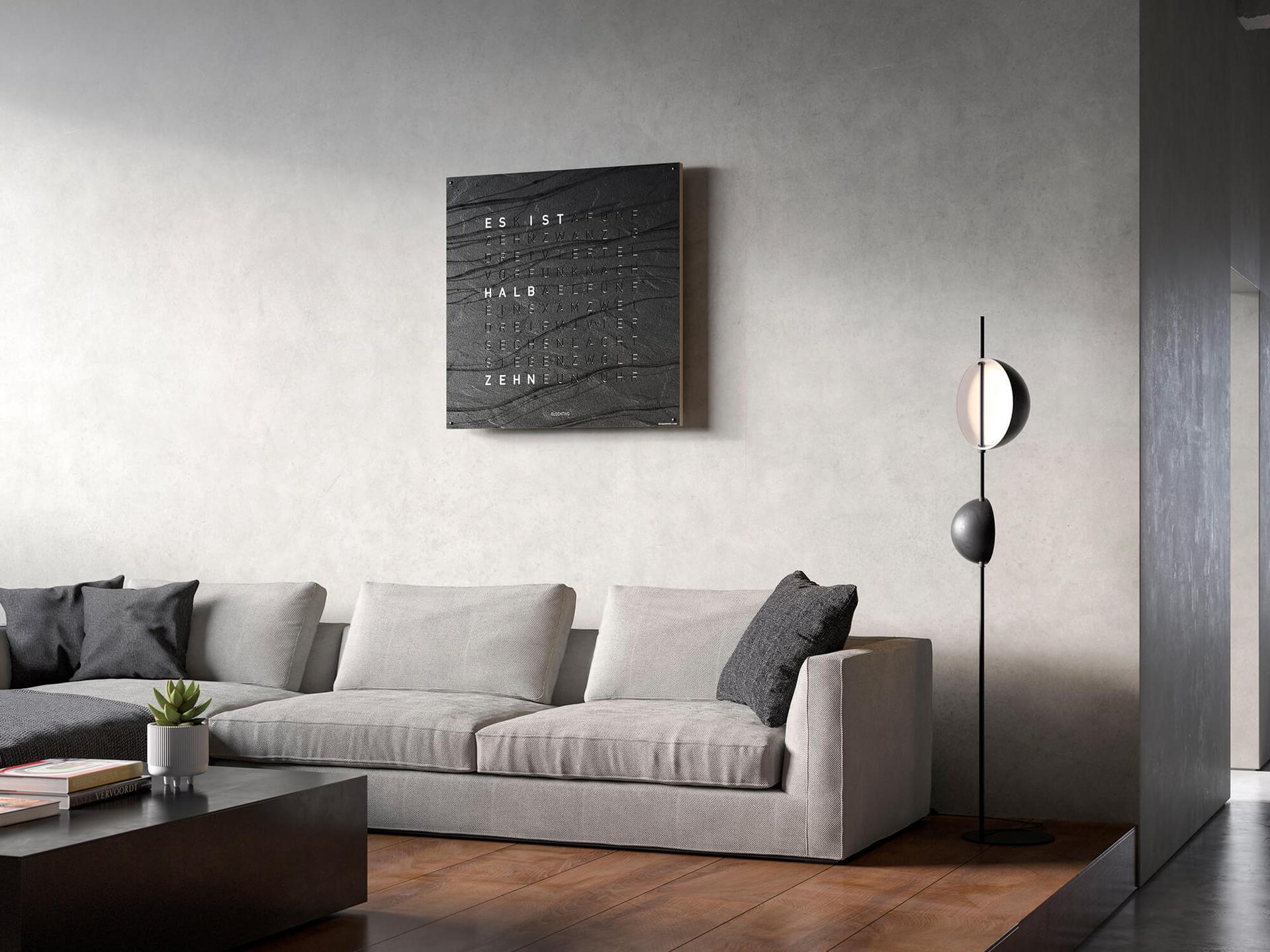 Qlocktwo Wanduhr in Schwarz mit Wortanzeige über hellem Sofa in neutralem Wohnraum
