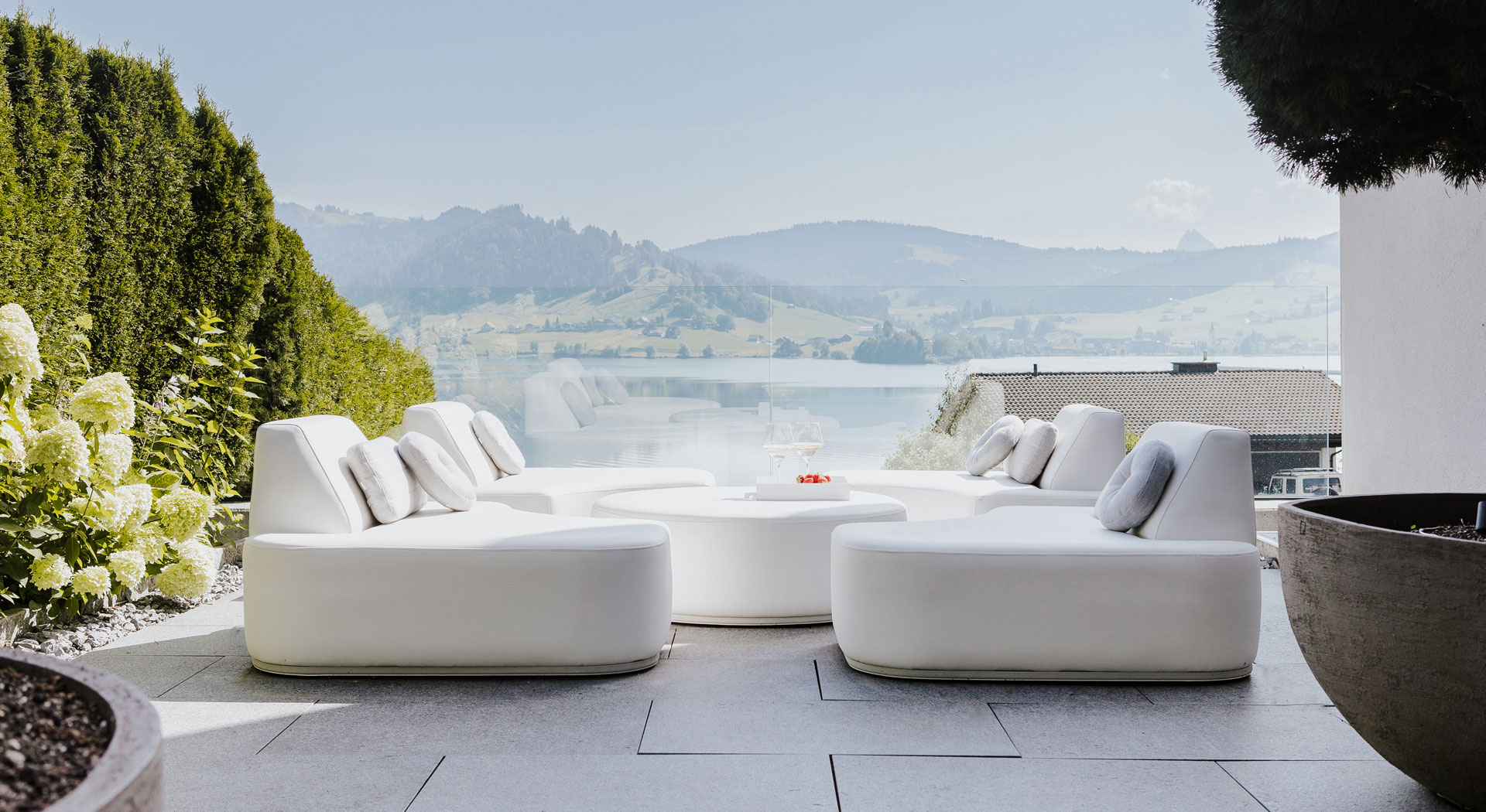 Weiße Outdoor-Lounge mit runden Modulen auf Natursteinboden, Panoramablick über See und Hügelkulisse.