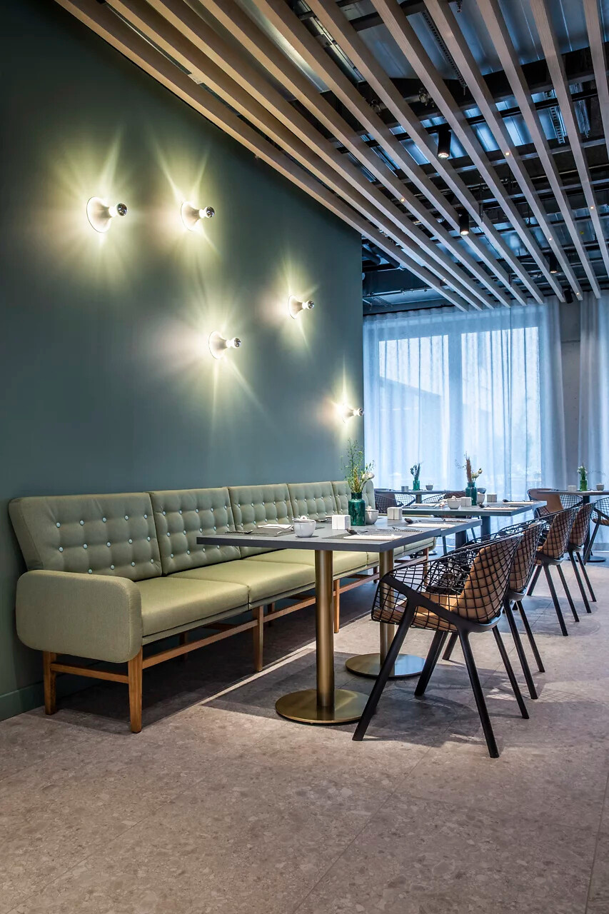 Grüne Polsterbank an Wand mit mehreren Mawa-Wandleuchten, Tisch und Stühlen im Vordergrund