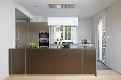 Die Einbauküche b3 von bulthaup besticht durch ihre zweifarbige Ausführung. Die Kücheninsel steht mit ihren bronzefarbenen Aluminiumfronten im Vordergrund, die lehmfarbene Hochschrankzeile nimmt sich dezent zurück. Kanten und Sockel der Küche sind in Aluminium gehalten. Eine hochglänzende Granitarbeitsplatte sorgt für spiegelnde Effekte. Die offene Wohnküche überzeugt mit harmonisch gestalteten Übergängen von Farben und Materialien. Hochwertige Einbaugeräte von Gaggenau und der höhenverstellbare und beleuchtbare Dunstabzug von Berbel sind weitere Highlights in diesem Küchentraum.